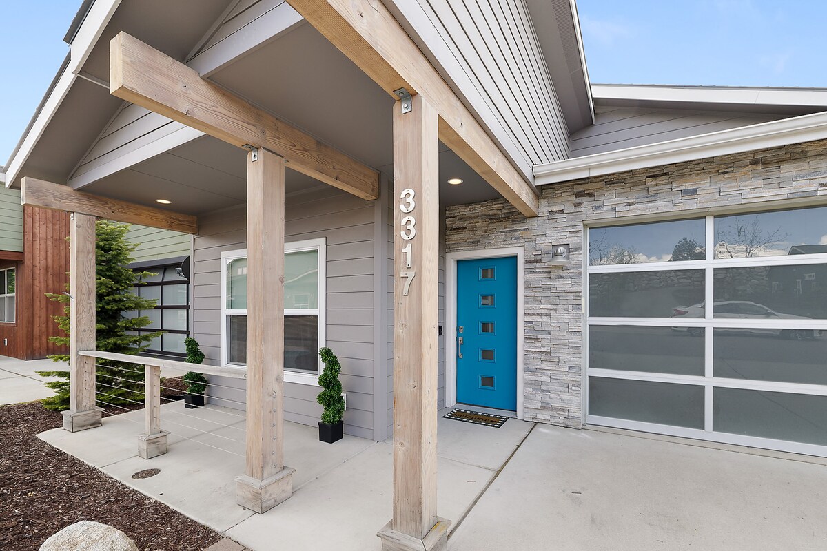 The entrance to the home features a modern design with a prominent blue front door and wooden beams creating a welcoming overhang. Flanking the door are two potted plants, and a smooth concrete pathway leads to the main entry.