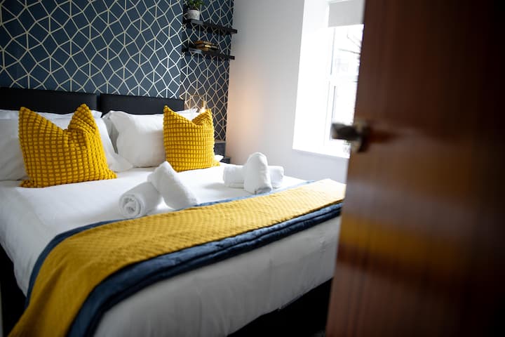 Светла и стилна двойна спалня с ярки жълти акценти, геометрични тапети и плюшени бели чаршафи за шикозно отдихване.