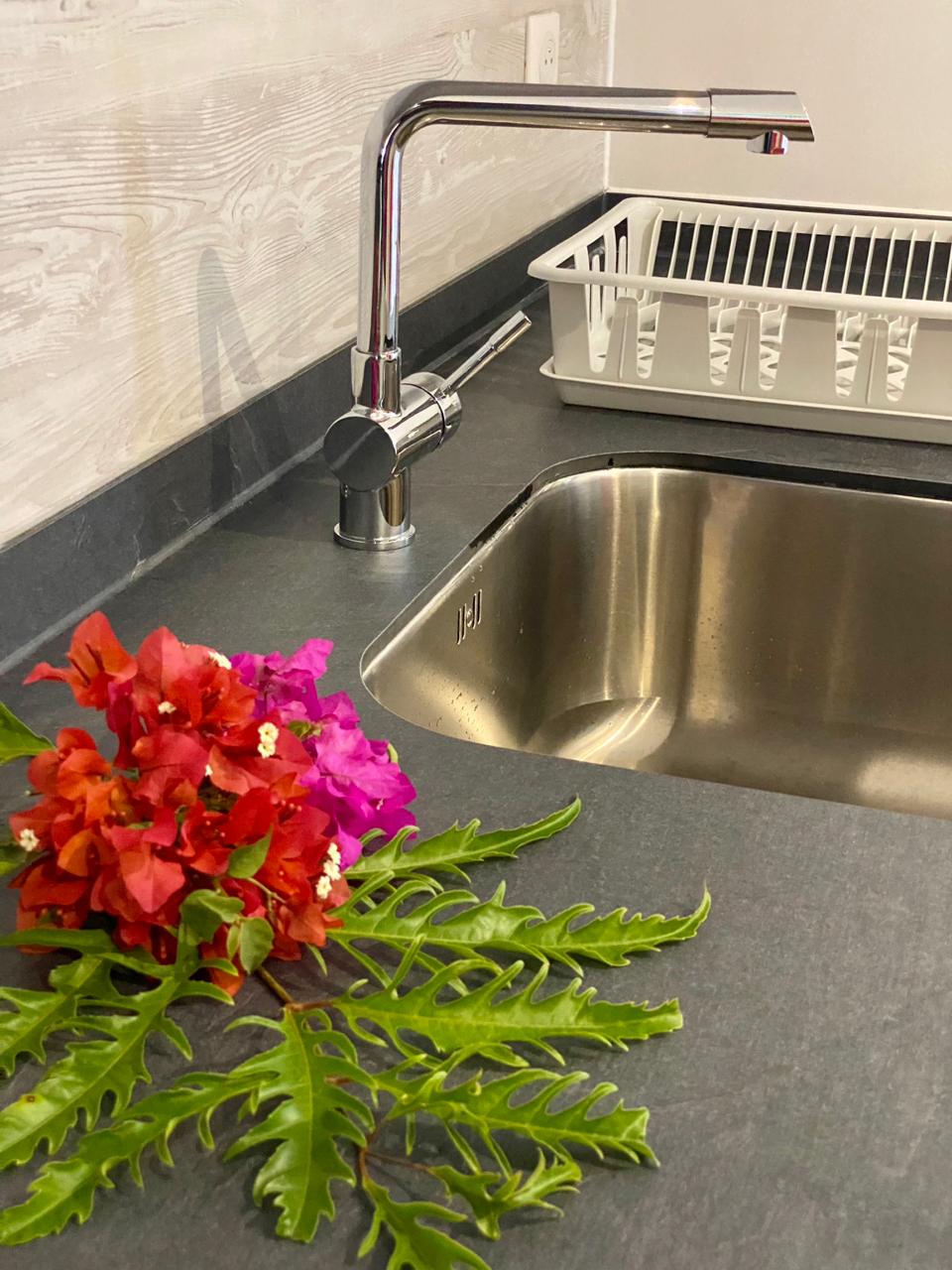 A kitchen countertop is shown, featuring a stainless steel sink and a sleek faucet. A white dish drying rack sits nearby. Brightly colored flowers and green foliage are arranged elegantly, adding a touch of nature to the functional space.