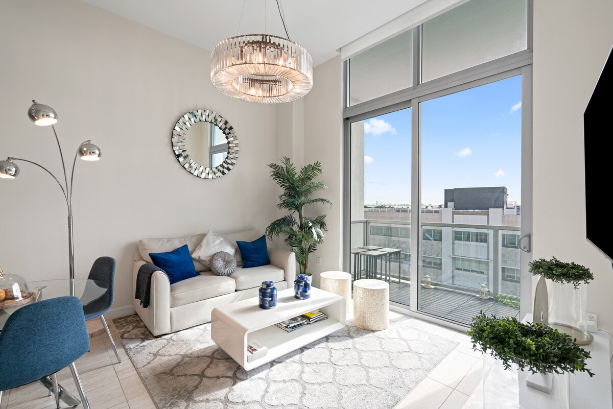 A modern living area features a light-colored sofa adorned with blue accent pillows. Large windows provide natural light and a view of the balcony. A sleek coffee table and decorative plants enhance the space. A round mirror and stylish chandelier add visual interest.