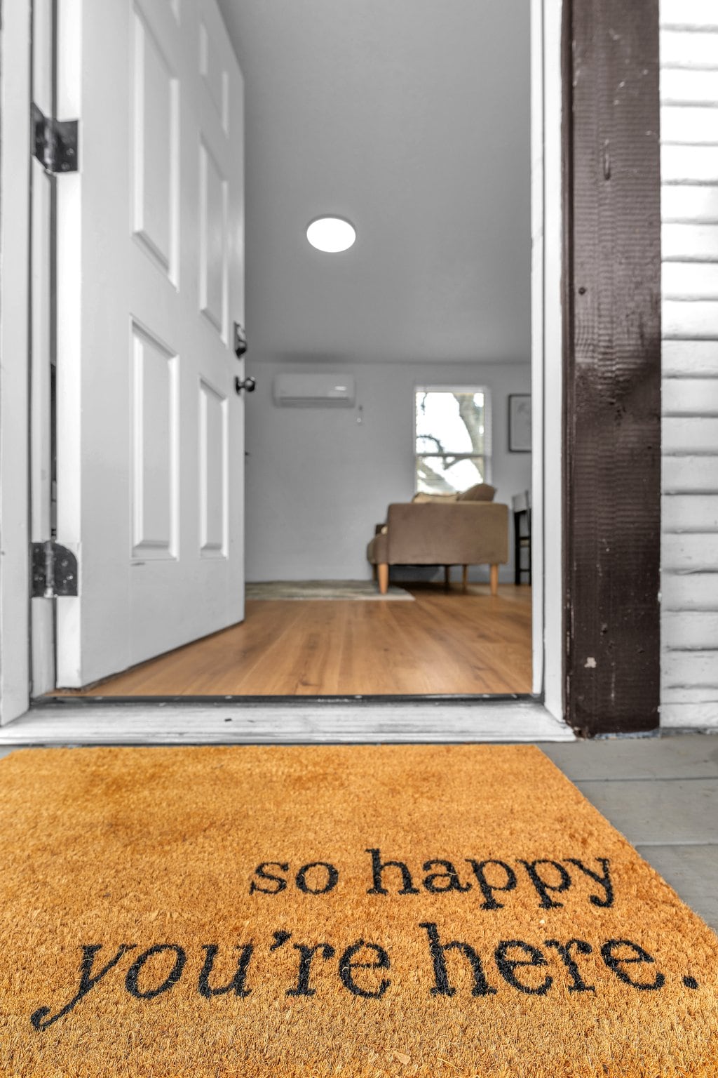 A welcoming entryway is highlighted by a doormat with the phrase 'so happy you’re here.' The open door reveals a glimpse of the interior, featuring light wooden flooring and a cozy living area visible in the background.