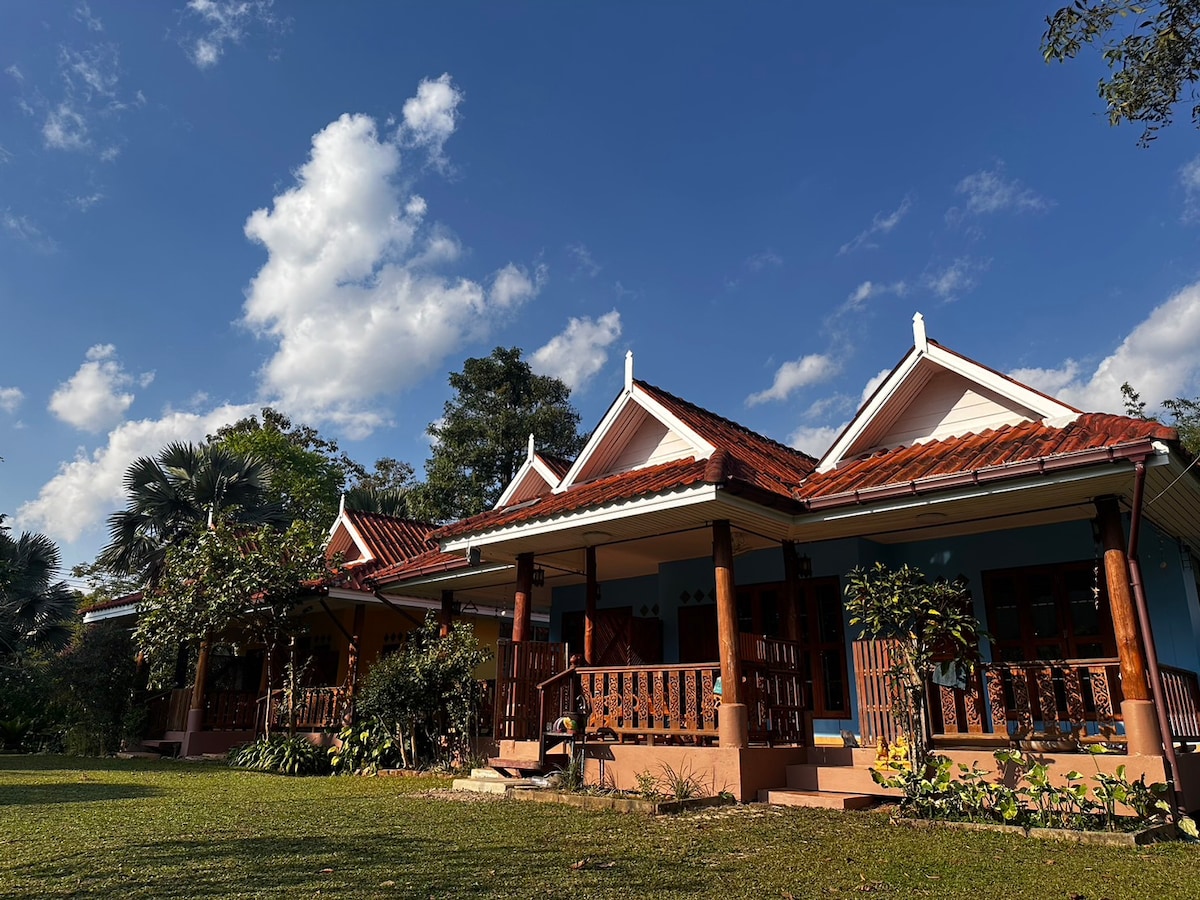 A serene view of multiple guesthouses is presented, featuring traditional architecture with sloped roofs and wooden balconies. Lush greenery surrounds the buildings, and a bright blue sky with scattered clouds adds to the tranquil atmosphere.