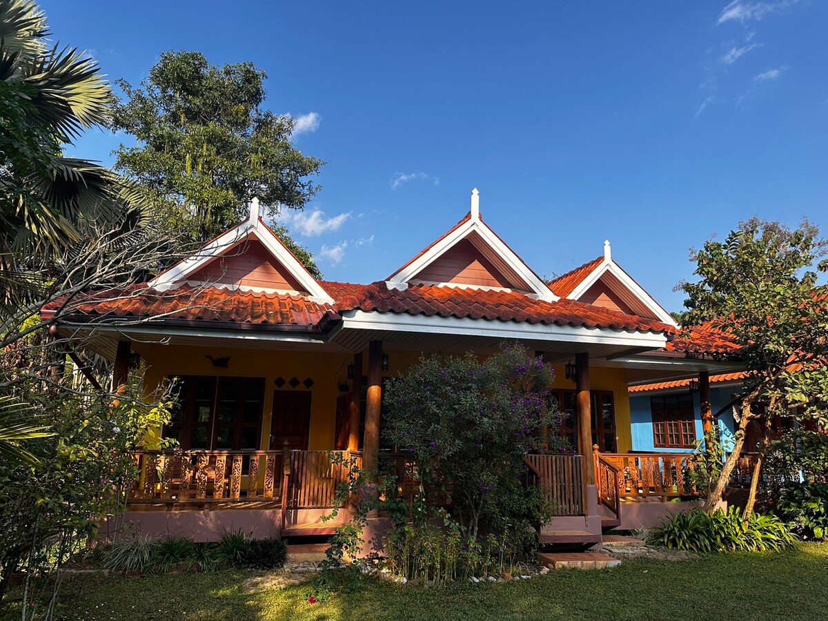 A charming guesthouse is set amidst lush greenery, featuring a vibrant orange facade complemented by a red tiled roof. The structure showcases multiple gabled roofs, with a welcoming porch adorned by wooden railings. Trees and shrubs surround the area, creating a serene outdoor atmosphere.