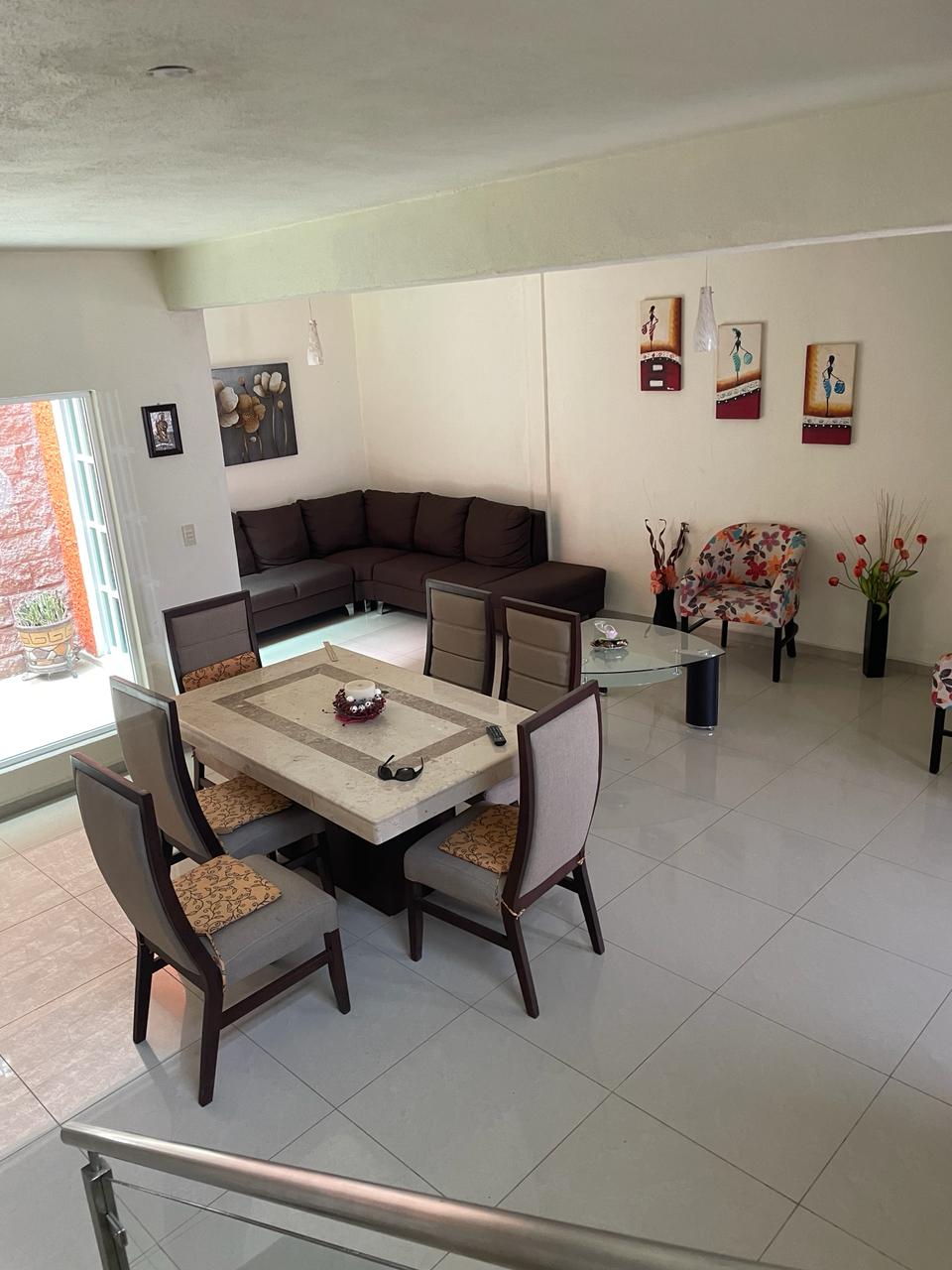 An open living space features a dark brown sofa accompanied by colorful chairs. A dining area with a large, rectangular table surrounded by six chairs is visible. Natural light enters through glass doors, highlighting the glossy tiled floor and decorative wall art.