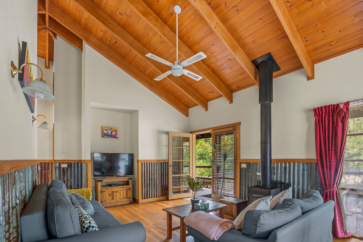 The spacious living area features high wooden ceilings, with a ceiling fan providing airflow. Two inviting sofas frame a central coffee table, while a wood-burning stove adds warmth. Natural light floods in through large windows with artistic window coverings, enhancing the cozy atmosphere.