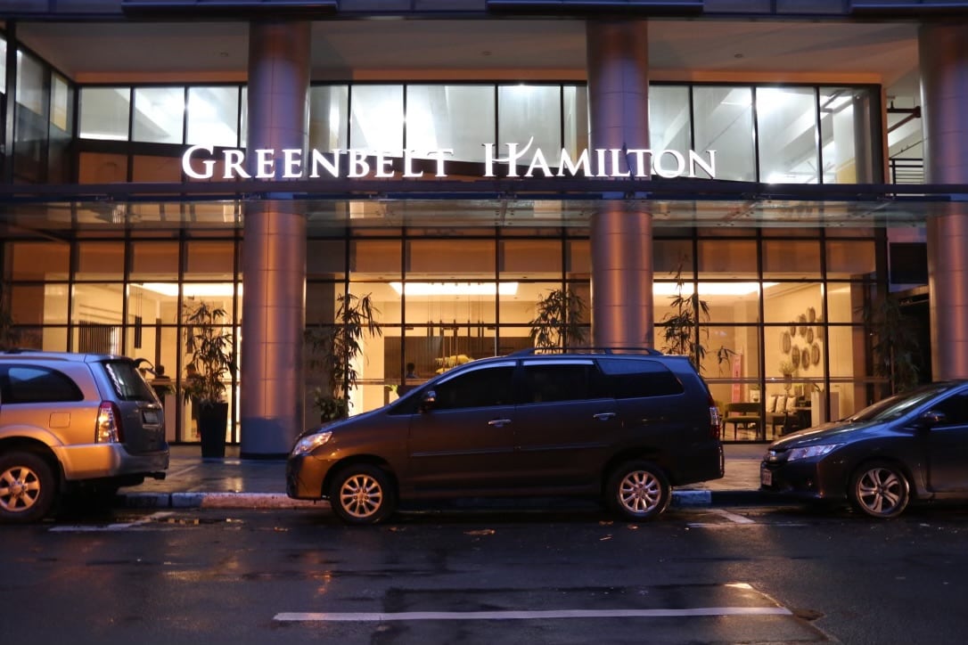 The entrance to Greenbelt Hamilton is showcased, featuring the building's name illuminated above large glass doors. The lobby area is visible through the glass, displaying a well-lit and inviting space. Several parked vehicles line the street, adding to the urban environment.