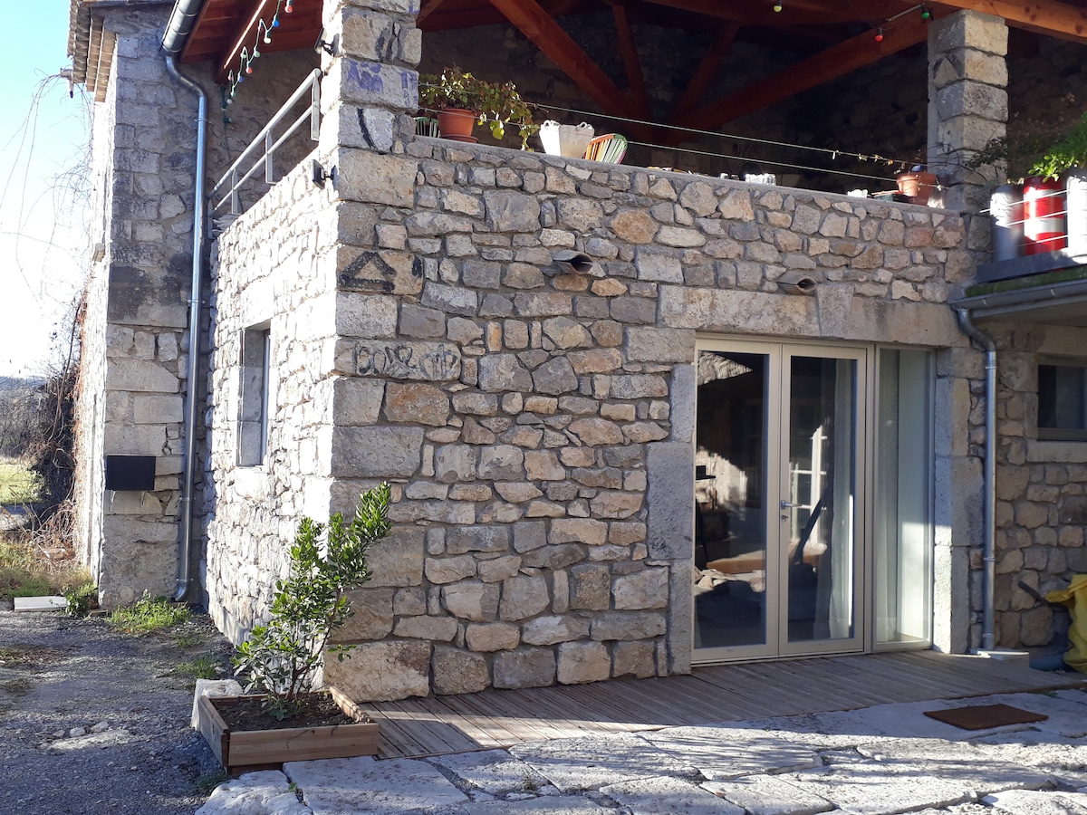 A rustic stone exterior features a covered area with wooden beams overhead. A glass door provides access to the interior, while flower pots are placed on a ledge. The surrounding space includes a wooden deck and elements of natural landscaping.