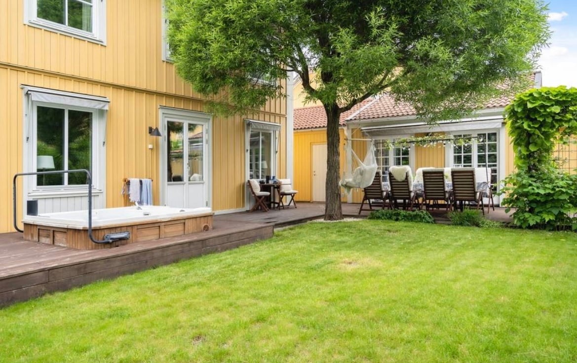 A charming outdoor space is presented, featuring a hot tub surrounded by wooden decking. The garden is adorned with a lush lawn and bordered by greenery. Comfortable lounge chairs are arranged around a dining table, offering a pleasant spot for relaxation and gatherings.