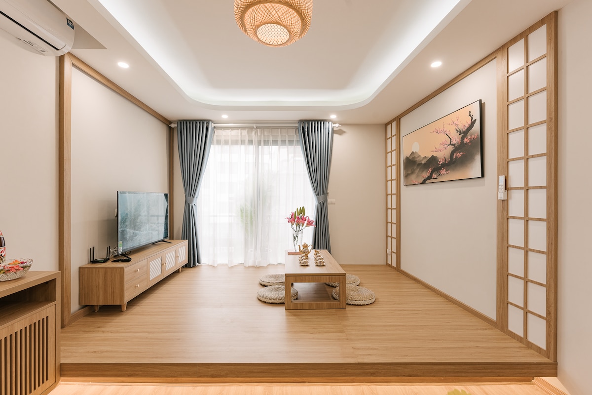 A spacious living area features a low wooden coffee table surrounded by circular cushions. A wall-mounted flat-screen TV is positioned opposite the large window, which is adorned with sheer curtains that softly diffuse natural light. Decorative elements include a wall art piece depicting a serene landscape.
