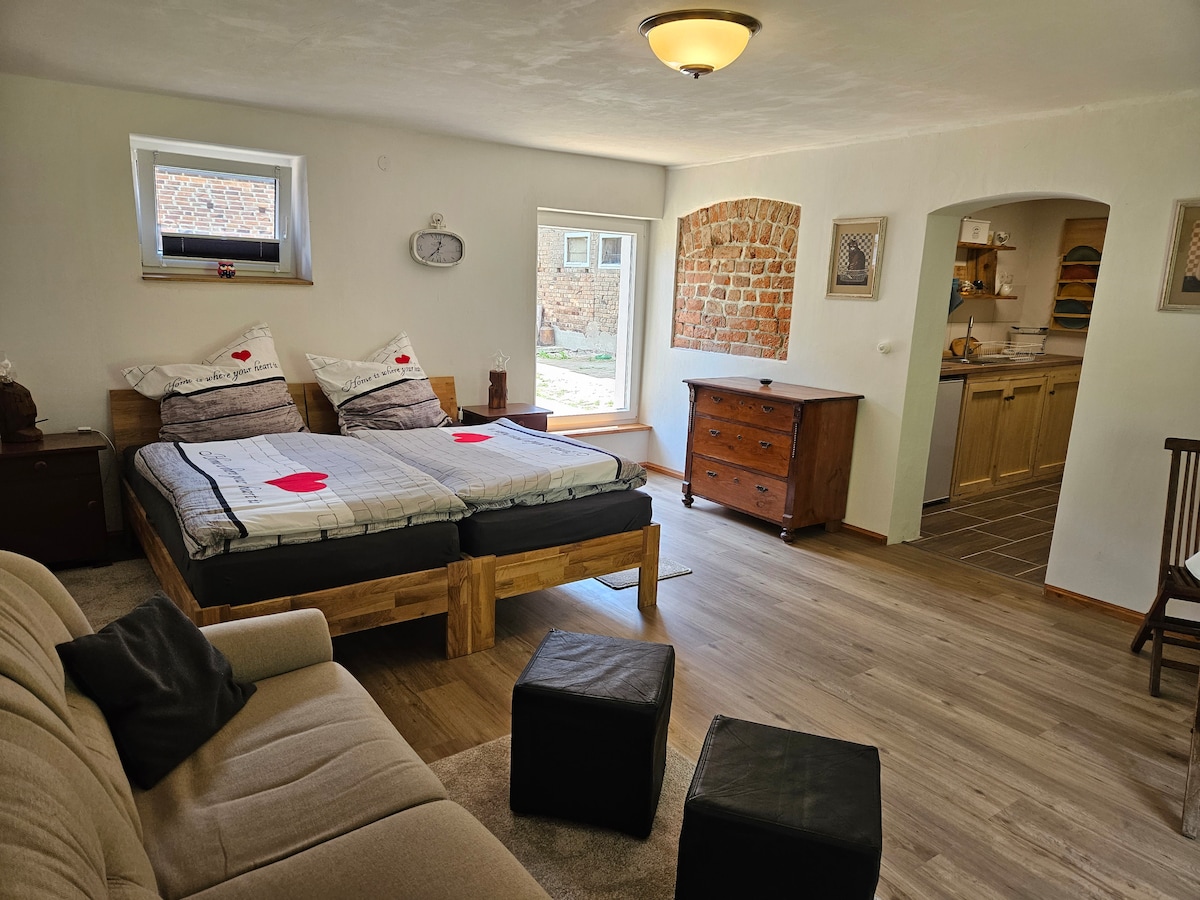 The spacious room features two twin beds with simple bedding, set against a neutral wall. A comfortable sofa is positioned nearby, with soft poufs on the floor. Natural light enters through a window, illuminating a rustic wooden dresser and an archway leading to the kitchen area.