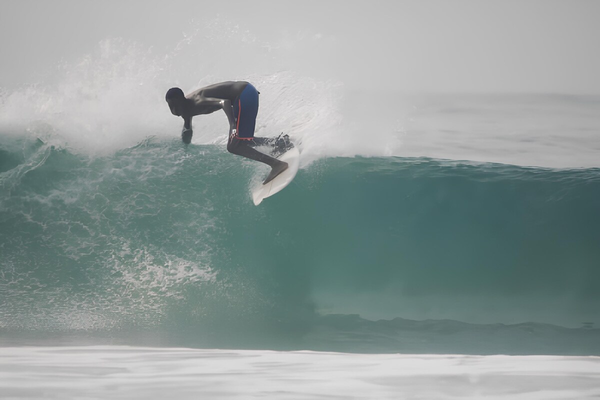 A surfer is captured riding a wave, with the ocean's water displaying shades of teal and white foam. The image highlights the dynamic motion of the wave as it breaks, creating a spray of water in the air.