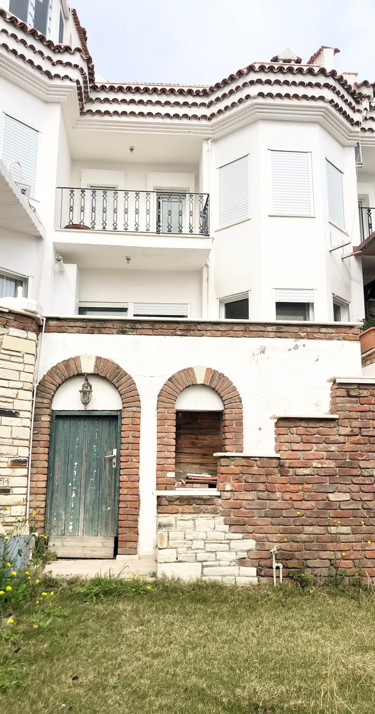 The exterior of a multi-level building features a combination of white walls and brick elements, with a decorative balcony above. A rustic green door and an arched stone structure are visible in the foreground, complemented by a grassy area with scattered flowers.