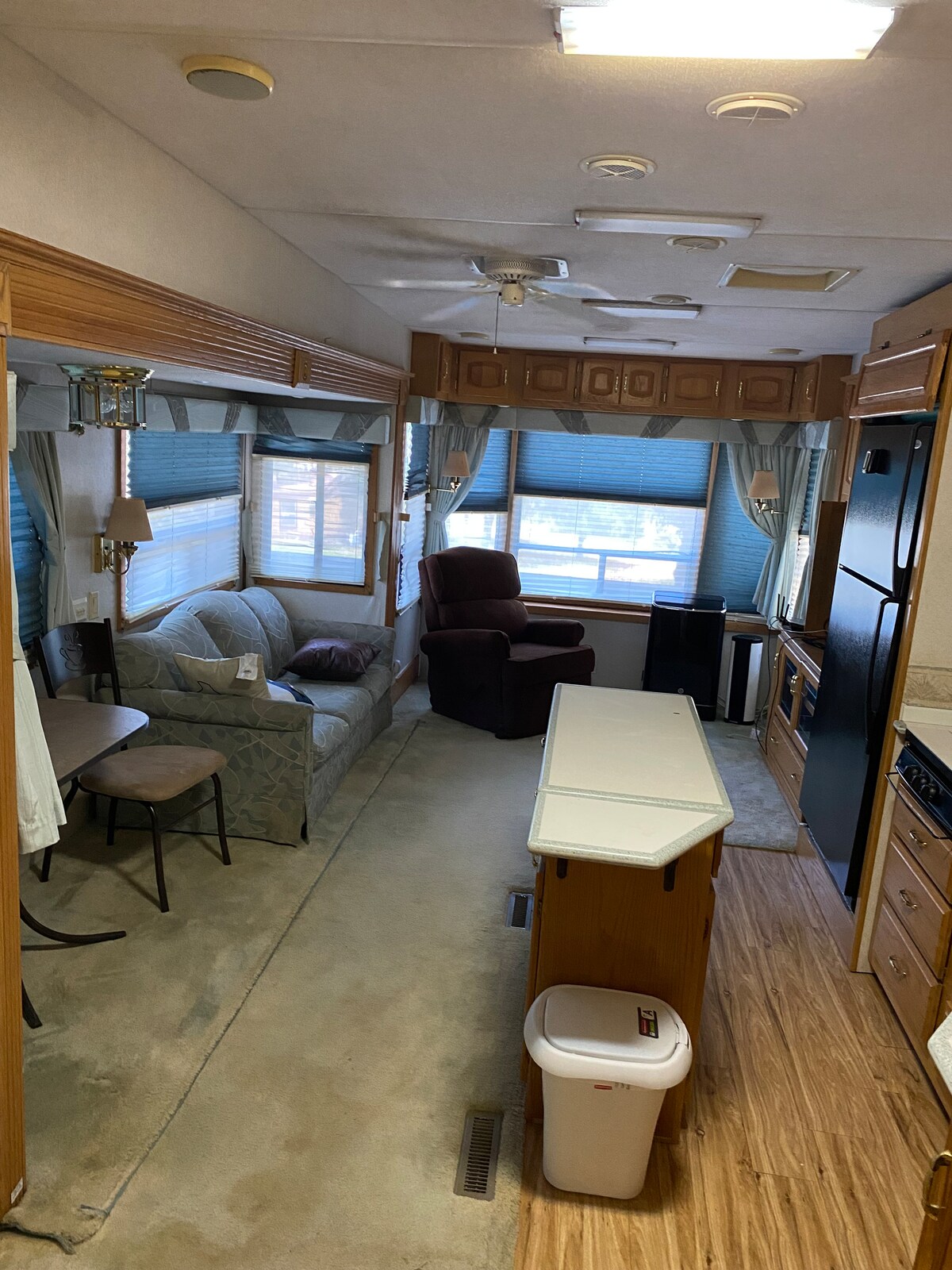 The interior of the RV is shown, featuring a cozy seating area with a sofa and an armchair beside a window. A small dining table with chairs is placed adjacent to the kitchen area, which includes cabinetry and appliances. Natural light enters through the blue-tinted window coverings.