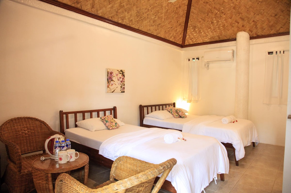 A serene sleeping area features three comfortable beds, each adorned with fresh linens. A woven chair and side table are positioned beside one bed, accompanied by a kettle and mugs. Natural light enters through two windows dressed with light curtains, highlighting the bamboo ceiling.