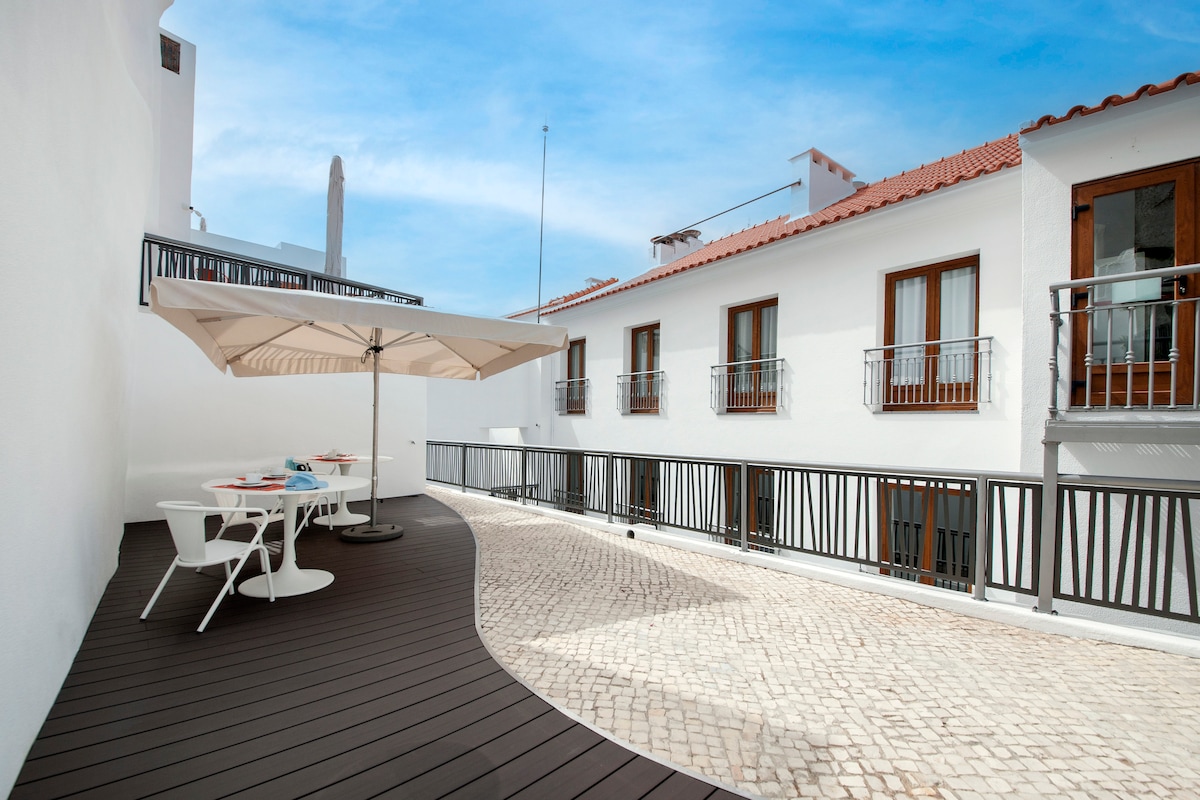 An outdoor terrace features a sleek wooden deck with a curved design. A large umbrella provides shade over a table set for dining. Surrounding the terrace are white walls and wooden-framed windows, offering a light and airy ambiance.