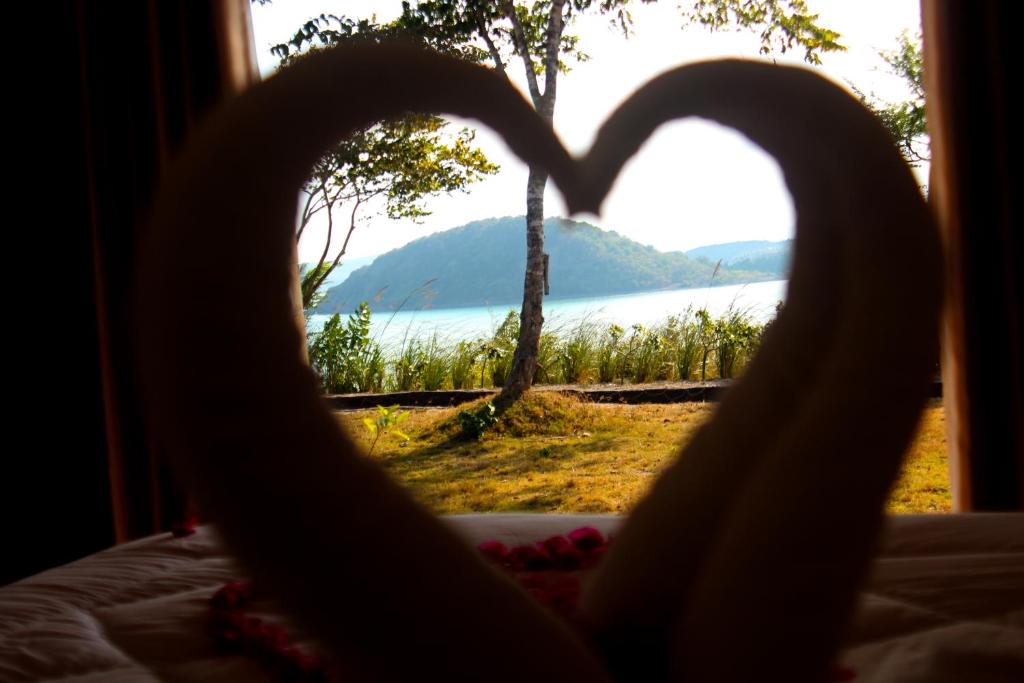 The image captures a scenic view from inside the room framed by towel art shaped like a heart. The background reveals a tranquil sea and a lush green island, with a gentle shoreline and trees visible in the foreground.