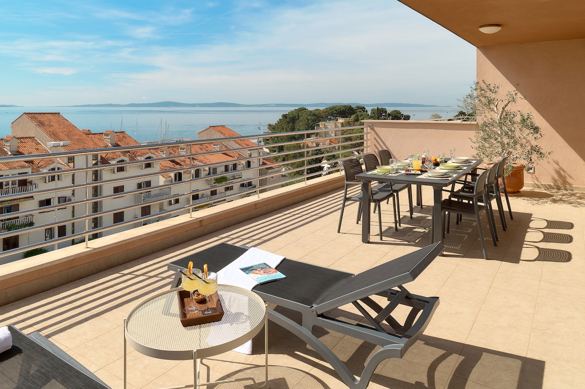 A spacious private terrace features modern outdoor furniture, including a dining table and loungers. The area is set against a backdrop of panoramic sea views and a sunny sky, inviting relaxation and enjoyment of coastal breezes.