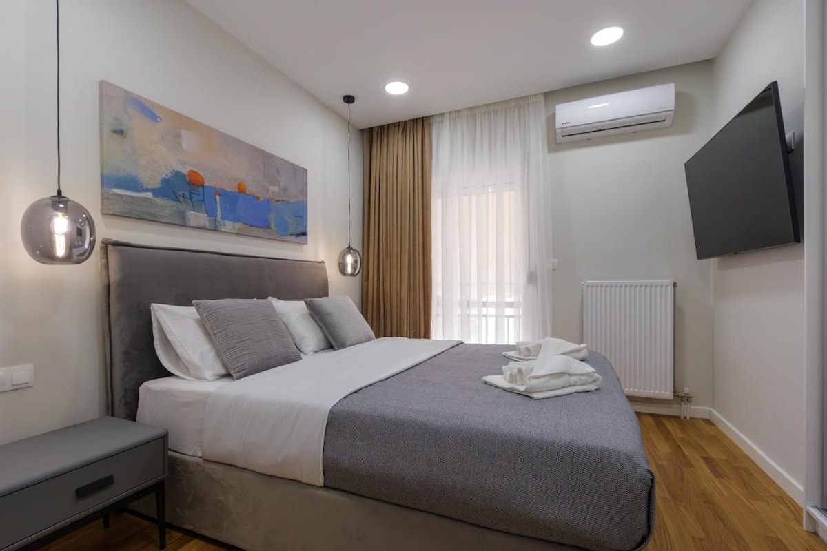 A master bedroom is featured with a spacious bed dressed in neutral linens and a soft gray throw. A large abstract painting adds a touch of color above the bed. Natural light filters through sheer curtains, while a flat-screen television is mounted on the wall.
