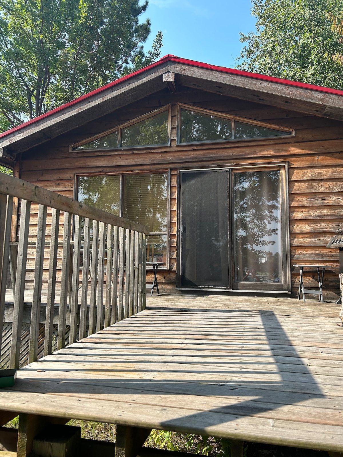 A rustic cabin exterior is shown, featuring a wooden deck leading to large glass doors. The façade is adorned with natural wood logs and a red roof. Surrounding trees provide shade, and several chairs can be seen on the deck.