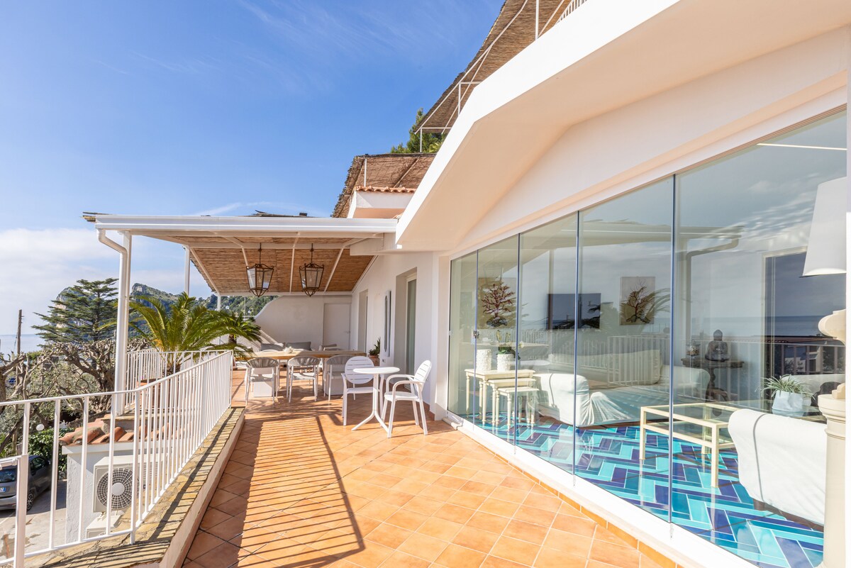 A spacious terrace is featured, adorned with a white table and chairs under a pergola. Floor-to-ceiling glass doors provide a view of the interior space, revealing airy living areas and inviting natural light. Lush greenery and panoramic sea views enhance the outdoor setting.