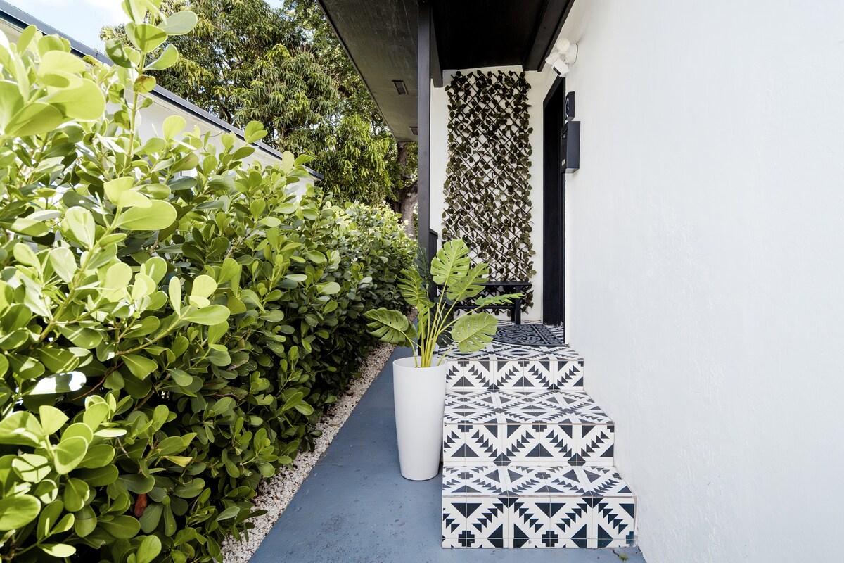 An outdoor entrance features patterned black and white tiles leading up three steps. Lush greenery flanks the pathway, with a tall white planter holding a leafy plant positioned to the side. The surrounding foliage adds a touch of nature to the entrance.