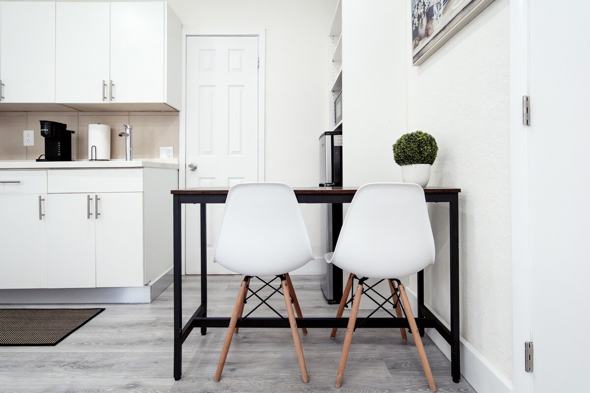 A kitchenette area is visible, featuring white cabinets and modern appliances. A dining table with two minimalist chairs is positioned in front of a window. A potted plant adds a touch of greenery to the space, contributing to a functional dining setup.