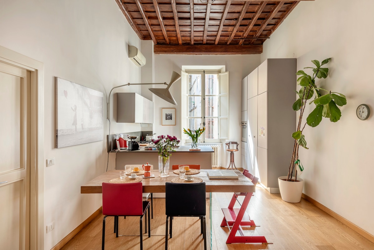 A spacious kitchen area features a large wooden dining table that seats six, adorned with plates and glasses. The light wooden flooring complements the exposed wooden ceiling beams. A large window allows natural light to fill the room, while potted greenery adds a touch of freshness.