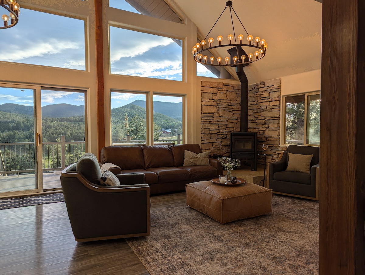 A welcoming living area features large windows that provide expansive views of the surrounding mountains. A comfortable leather sofa and chairs are arranged around a round coffee table, with a cozy rug underneath. A stone fireplace adds a rustic touch to the room.