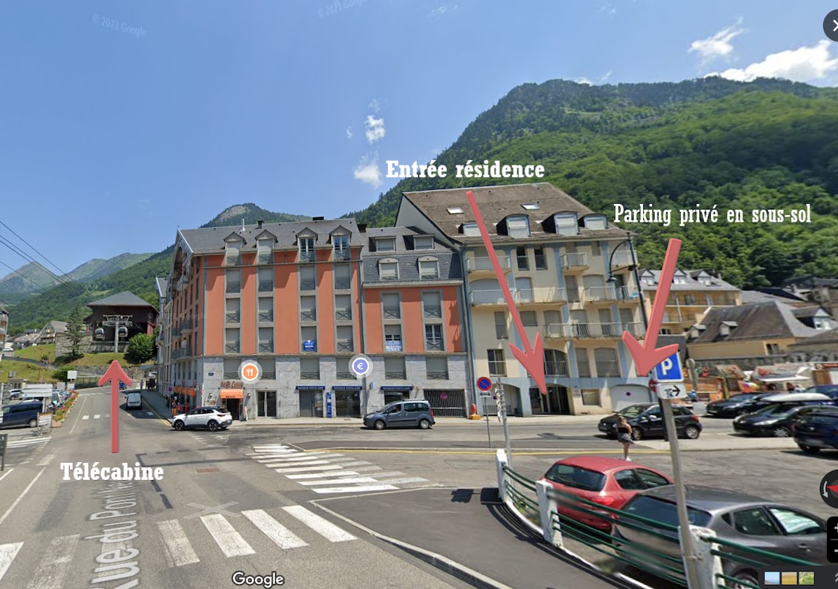 A view of the apartment building's exterior is displayed, showcasing a multi-story structure. Signs indicate the entrance to the residence, as well as nearby parking options. The surrounding landscape features mountains, highlighting the location's natural beauty.
