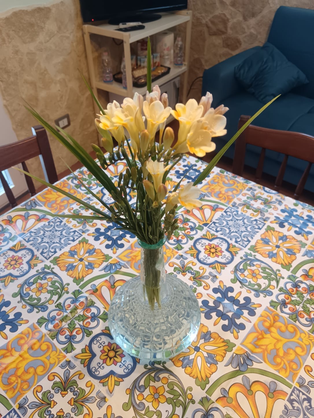 A clear glass vase holds an arrangement of yellow flowers, complemented by green leaves. The vase is placed on a patterned tablecloth featuring vibrant colors and intricate designs. In the background, a cozy seating area and television are subtly visible.