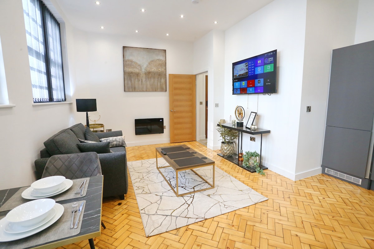 An open-plan living space is displayed, featuring a comfortable seating area with grey sofas and a modern coffee table. A large flat-screen TV is mounted on the wall, and warm lighting enhances the inviting ambiance. A stylish patterned rug complements the wooden flooring.