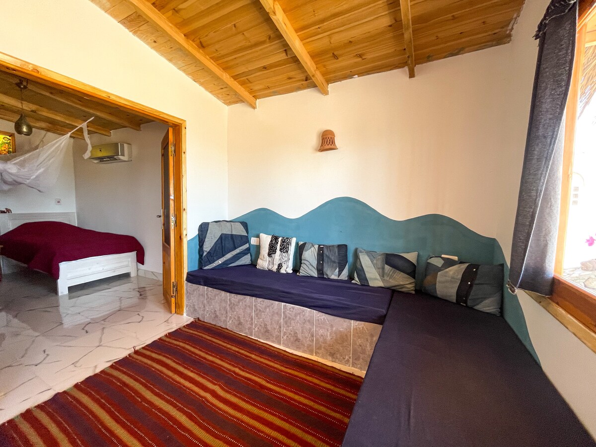 A seating area is designed with a wave-patterned backrest and various cushions in neutral tones, positioned against a wooden ceiling. A doorway leads to a separate sleeping space, while light enters through a large window adorned with simple curtains.