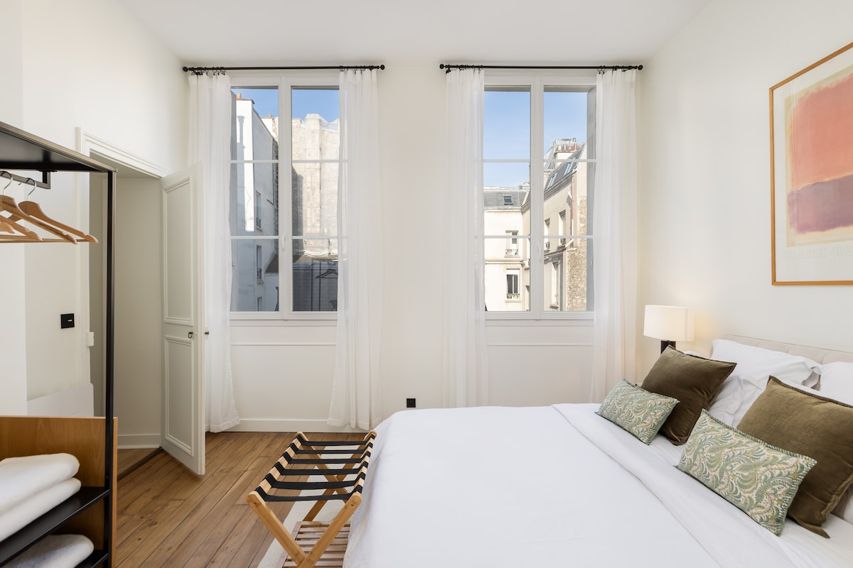 The main bedroom features a spacious bed adorned with decorative pillows. Light-colored walls and large windows allow natural light to illuminate the room. A simple wooden clothing rack is positioned beside the bed, adding to the minimalist decor.