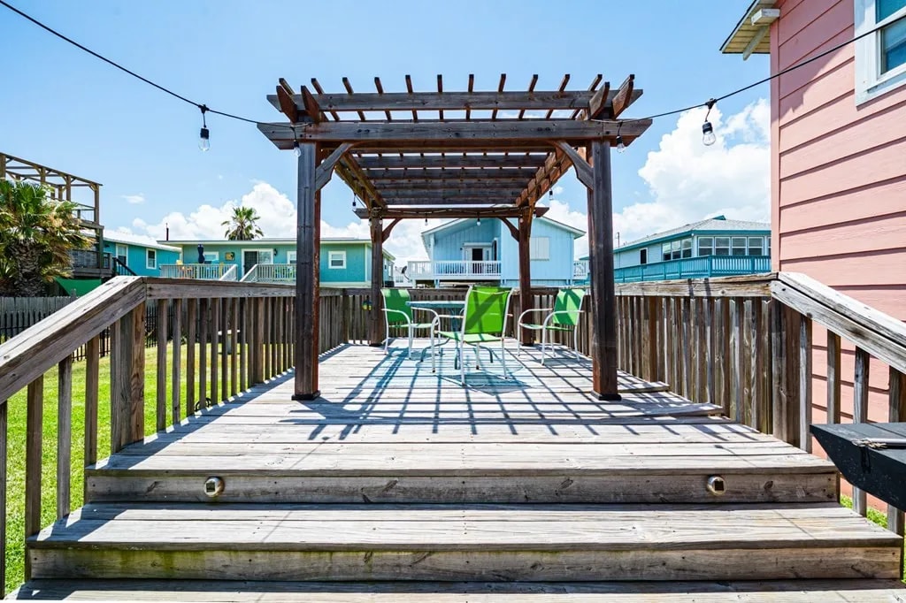 A spacious deck area is featured, offering a wooden pergola supported by posts. Four bright green chairs are arranged beneath the structure, utilizing natural light from the open sky. Surrounding greenery and nearby buildings are visible in the background.