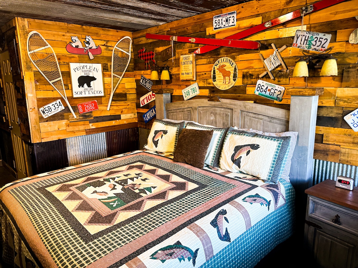 A bedroom is designed with a rustic theme, featuring a spacious bed adorned with a colorful quilt showcasing fish patterns. The wooden walls are decorated with vintage license plates and fishing gear. Soft lighting is provided by bedside lamps mounted on either side.