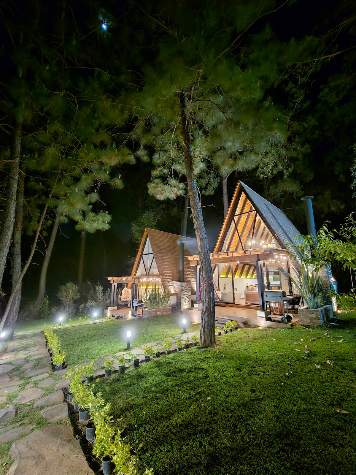 The exterior of the cabin is illuminated at night, showcasing an A-frame design nestled among tall trees. A well-maintained garden with pathway stones and subtle lighting enhances the serene ambiance. A cozy seating area is visible on the porch, inviting relaxation in nature.