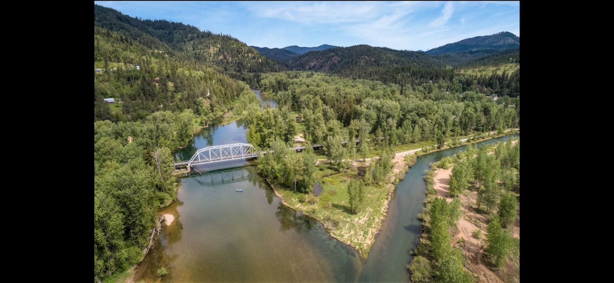 An aerial view captures the winding Coeur d'Alene River, bordered by lush greenery and gently rolling hills. A modern bridge spans the river, while calm waters reflect the surrounding trees and sky. The landscape invites exploration, with various paths visible along the riverbanks.