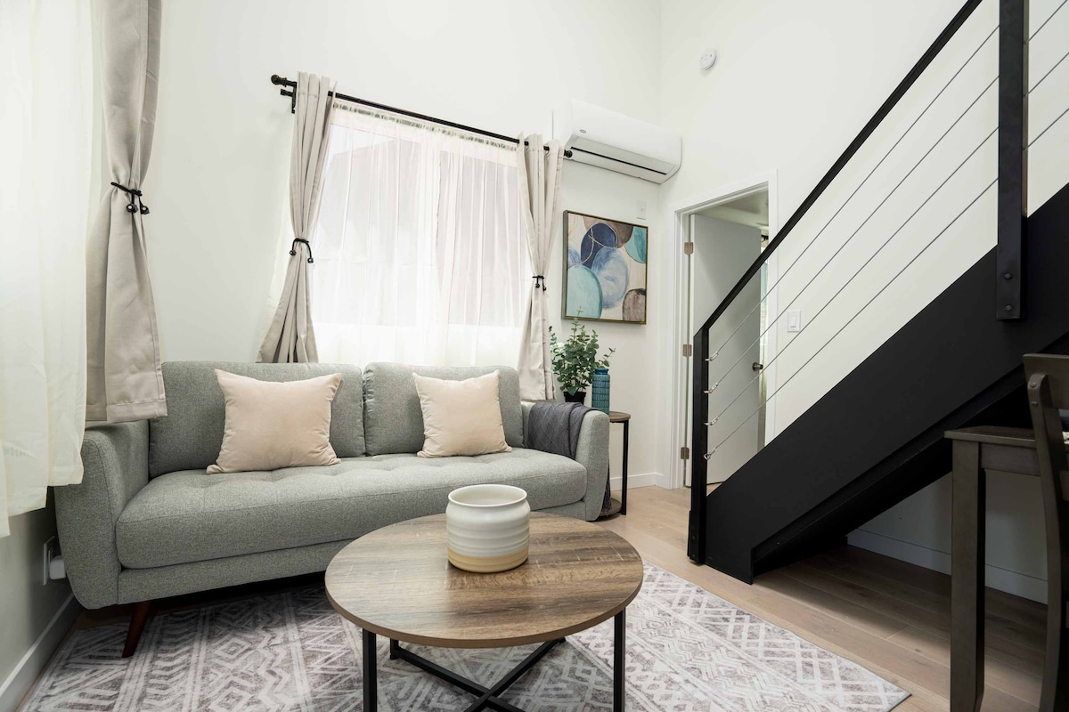 A cozy living area features a soft gray sofa adorned with light-colored pillows, positioned beside a round coffee table. A large window allows natural light to fill the space, complemented by sheer curtains. An artwork and lush plant add visual interest, while a staircase leads to the upper level.