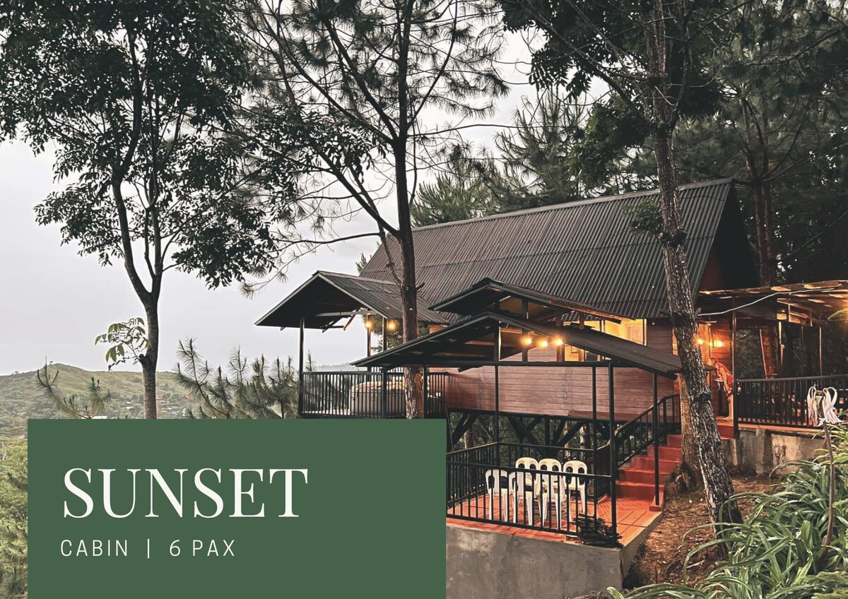 The exterior of the Sunset Cabin is presented, showcasing a two-story structure surrounded by tall trees. Warm lights illuminate the balcony area, where outdoor seating can be seen. The sloped roof complements the natural landscape, creating a harmonious blend with the environment.