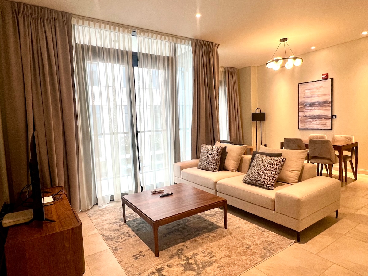 A spacious living area features a neutral-colored sofa adorned with decorative pillows. Natural light streams through tall, sheer curtains over expansive glass doors. A wooden coffee table sits at the center, accompanied by a dining table and modern dining chairs in the background.