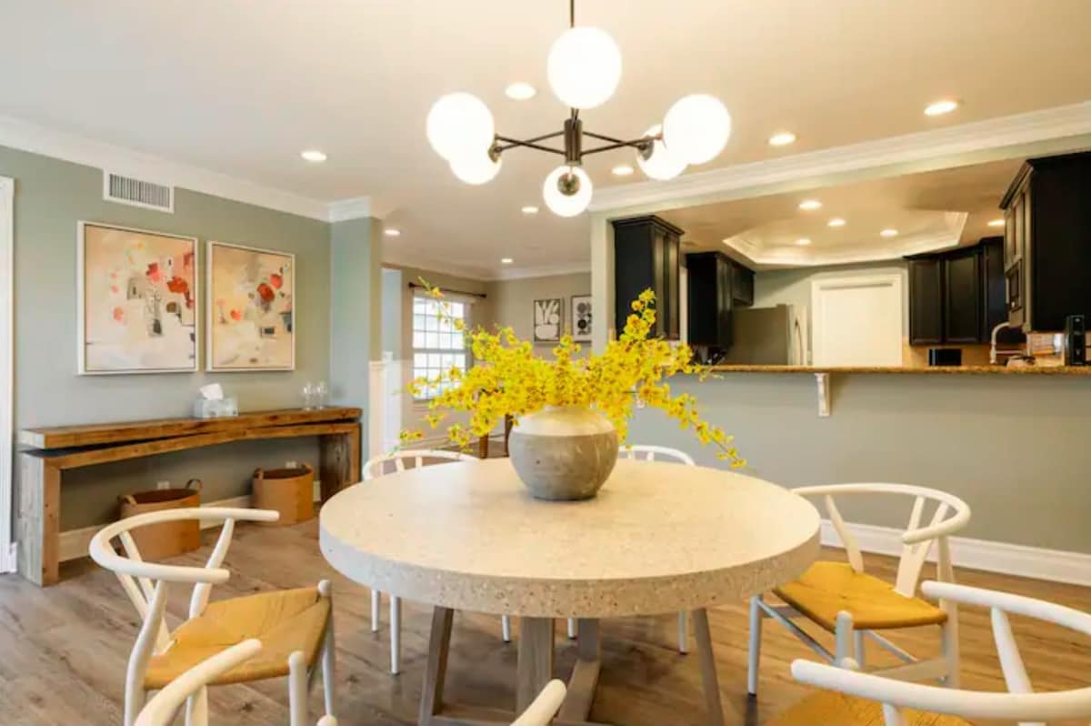 A round dining table is centered in a light-filled area, surrounded by four wooden chairs with woven seats. A large decorative vase filled with yellow flowers is placed on the table, adding a touch of color. The background features an open kitchen with dark cabinetry.