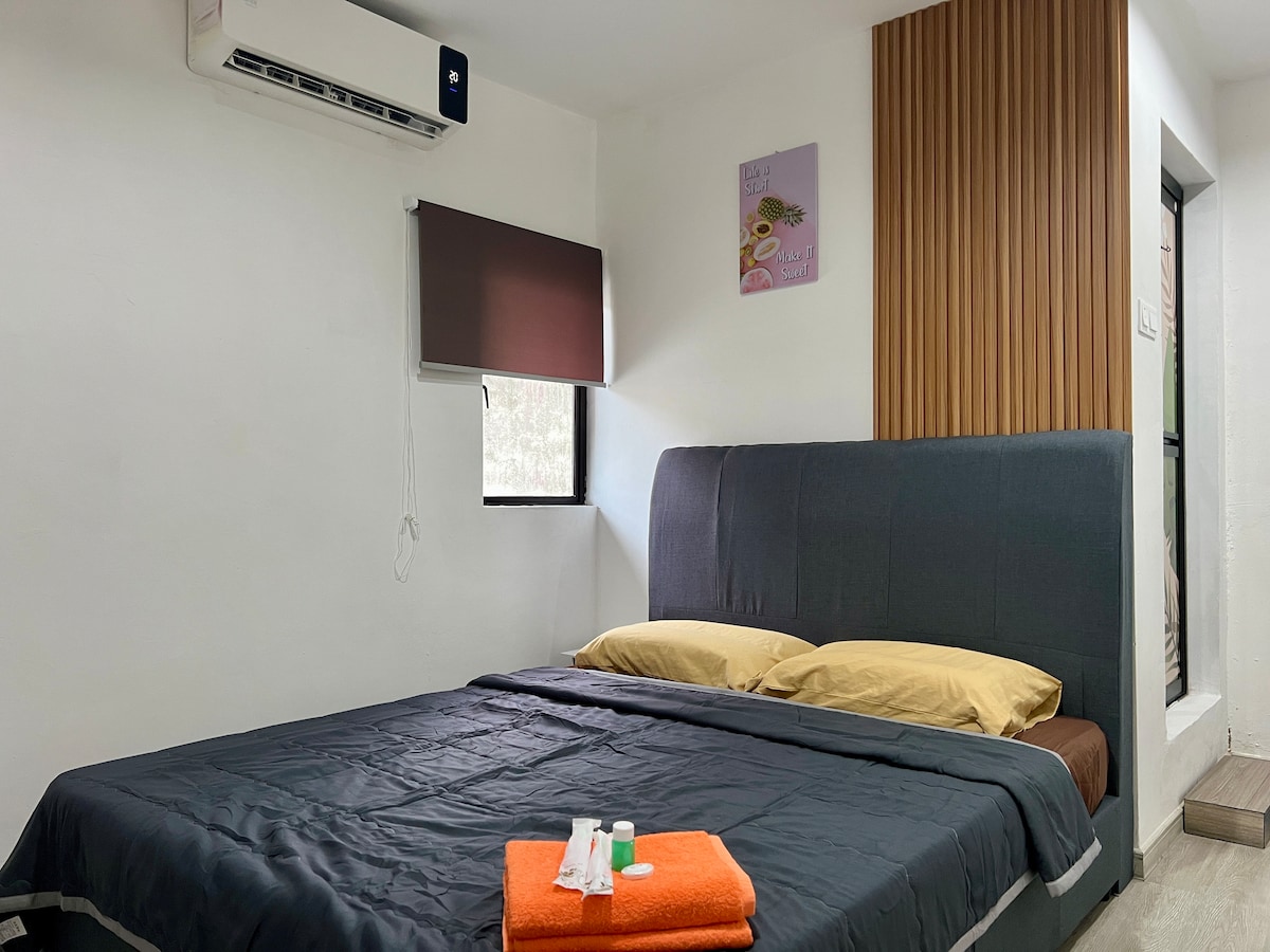 A spacious sleeping area is furnished with a dark gray bedspread atop a large bed, complemented by two golden-yellow pillows. An orange towel is neatly folded on the bed, accompanied by small toiletries. Natural light enters through a window with a brown window shade, enhancing the minimalist decor.