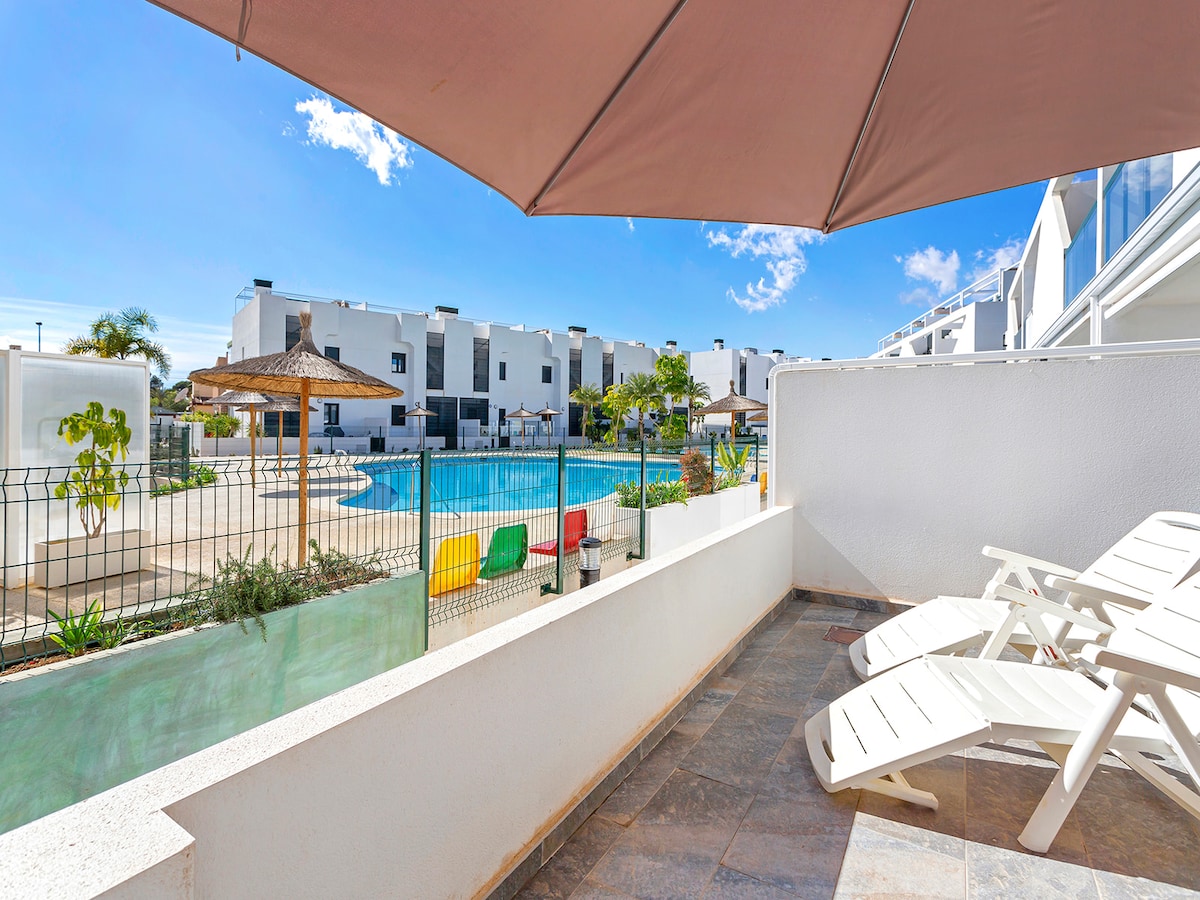 A terrace area is showcased with several lounge chairs under a large umbrella, overlooking a swimming pool. The pool is surrounded by greenery and colorful seating options. The modern residential buildings are visible in the background against a clear blue sky.