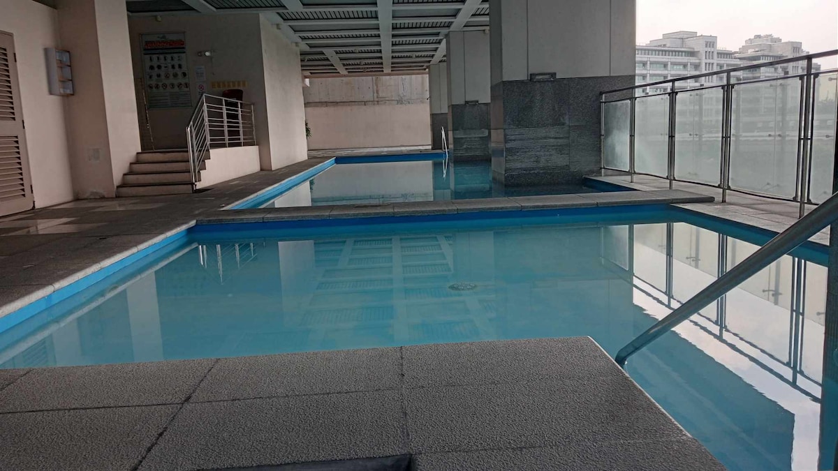 The outdoor swimming pool is framed by a spacious deck and glass barriers, offering a clear view of the surrounding area. The water appears inviting, reflecting the sky above, and a set of stairs leads to a poolside area for relaxation.