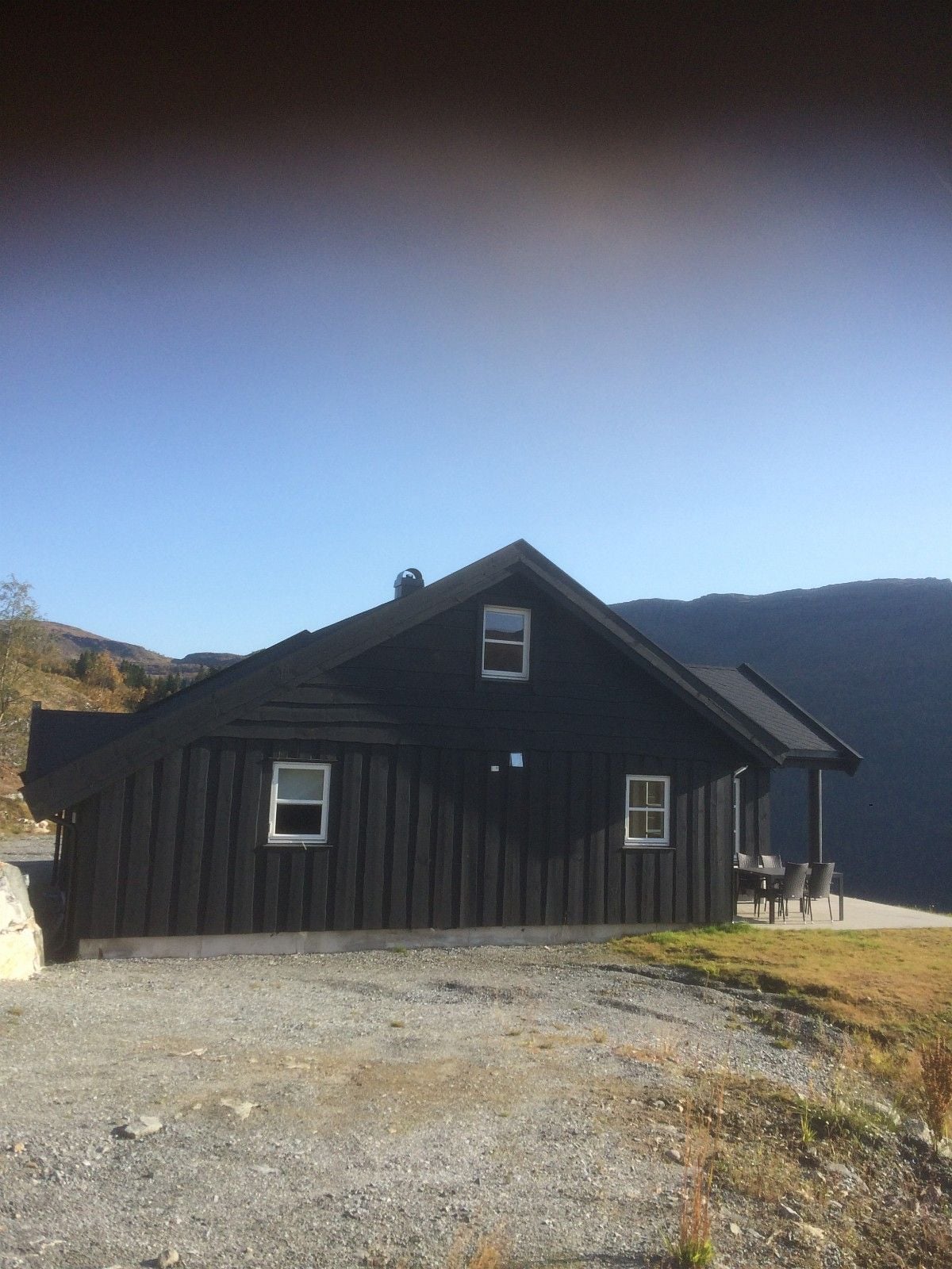 The side view of a dark wooden cabin is shown against a clear blue sky. The structure features a peaked roof and multiple windows. A small outdoor seating area is visible, positioned on a gravel path that leads to a scenic mountain backdrop.
