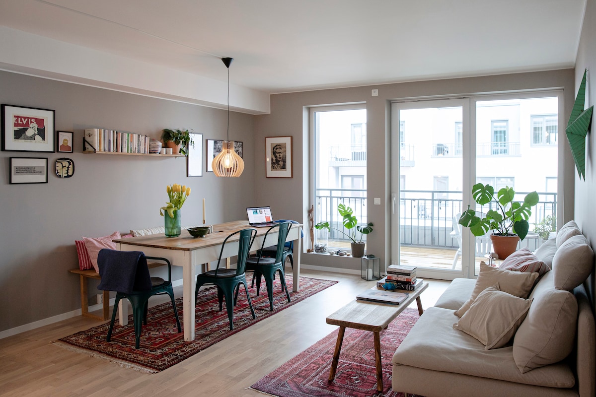 A spacious living and dining area features a light wood dining table surrounded by teal chairs. Natural light floods in through large glass doors that lead to a terrace. A comfortable sofa is placed nearby, complemented by decorative plants and artwork on the walls.