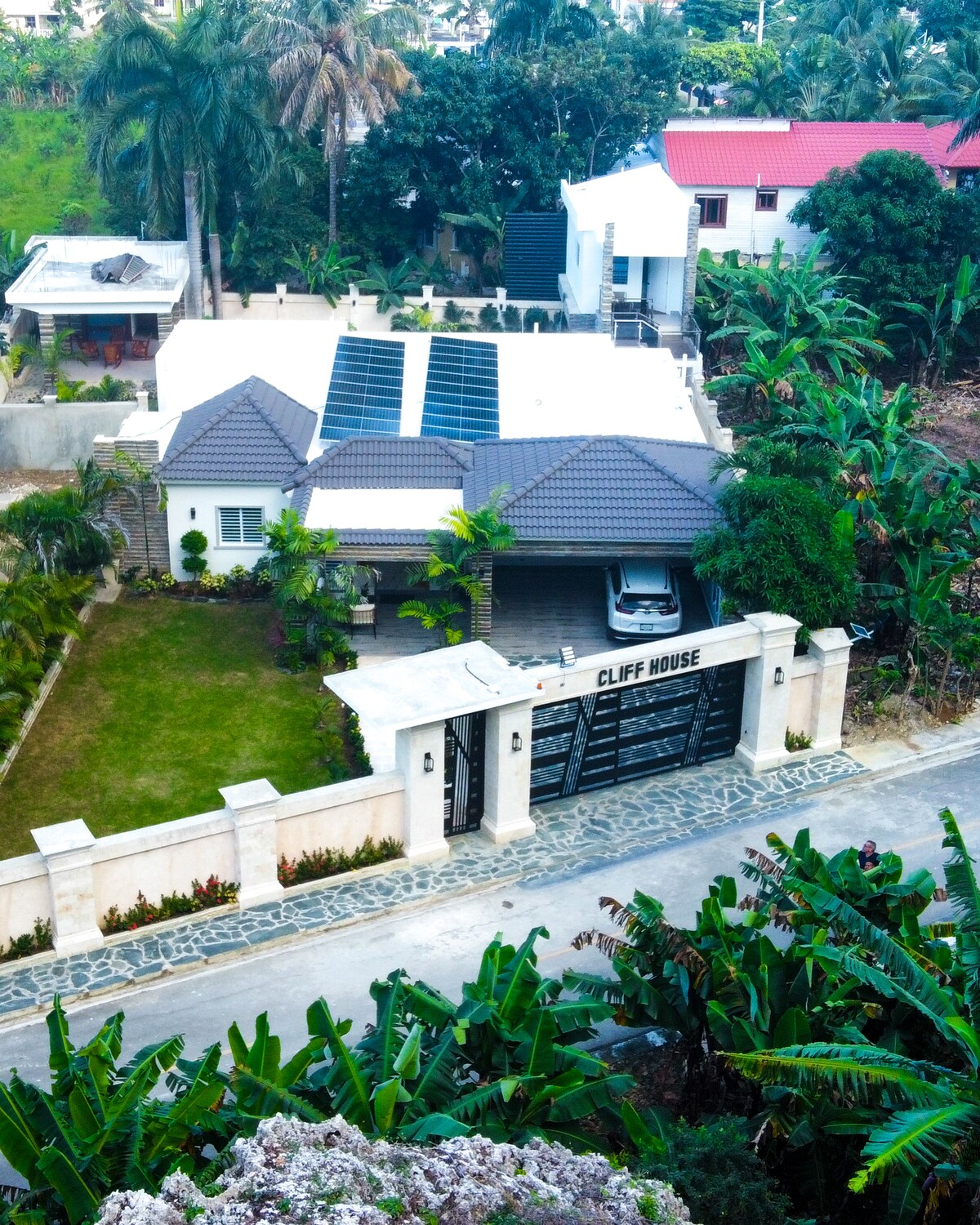 The exterior of a modern house is showcased, featuring a well-maintained lawn and tropical landscaping. Solar panels are visible on the roof. A gated entrance marked 'CLIFF HOUSE' leads to a spacious driveway with a parked vehicle. Surrounding greenery adds to the serene environment.