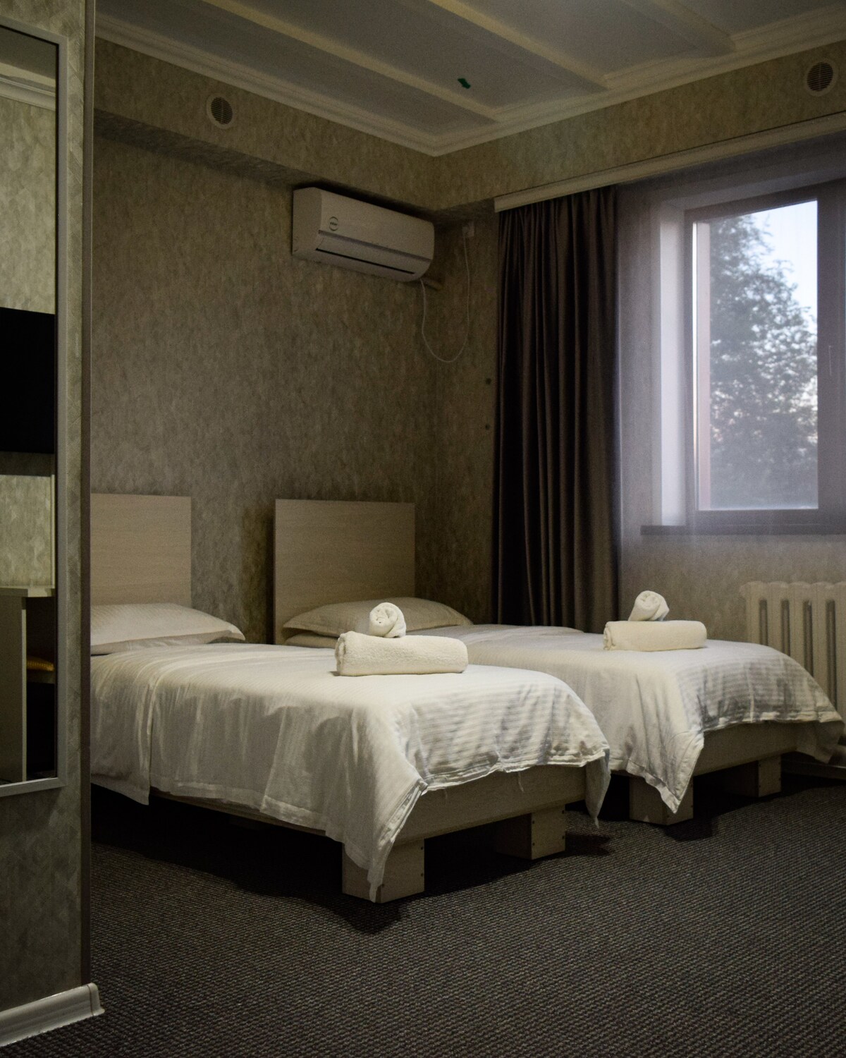 A well-lit guest room features two spacious beds with white linens and rolled towels on top. A large window allows natural light to fill the space, accompanied by soft curtains. An air conditioning unit is mounted above, and the neutral-toned walls add a calming atmosphere.