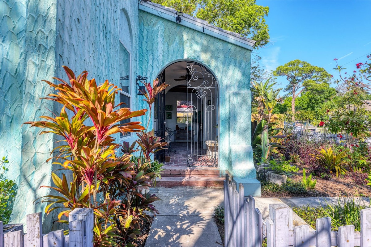 A charming entrance is framed by a light blue archway, leading to an inviting interior. Lush tropical plants with vibrant foliage decorate the path, enhancing the serene atmosphere. The exterior features textured walls, while the inviting garden showcases diverse native flora.