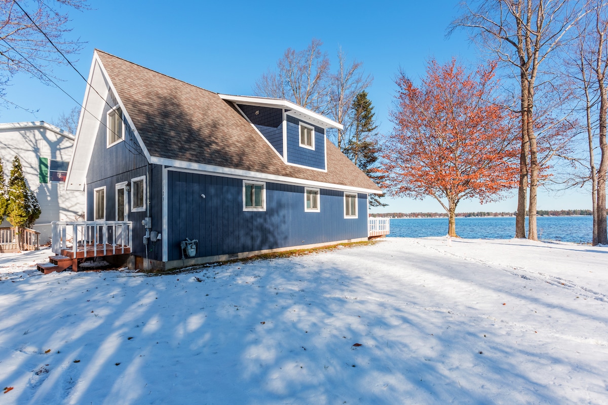 A blue, two-story cottage is set against a backdrop of winter scenery. The structure is surrounded by snow-covered ground and features a large tree with red foliage. Views of the lake can be seen in the distance.